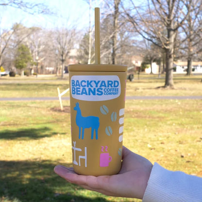 Yellow tumbler with 'Backyard Beans Coffee Company' branding held in a hand outdoors.
