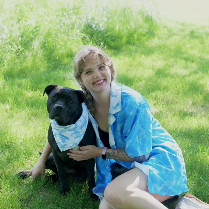 Woman in a blue llama button up sitting on grass with a black dog wearing a llama bandana.