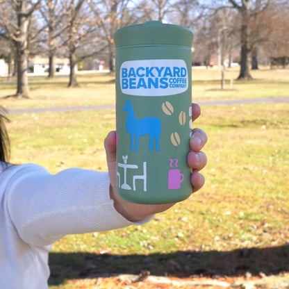 Person holding a green tumbler with 'Backyard Beans Coffee Company' branding in a park setting.