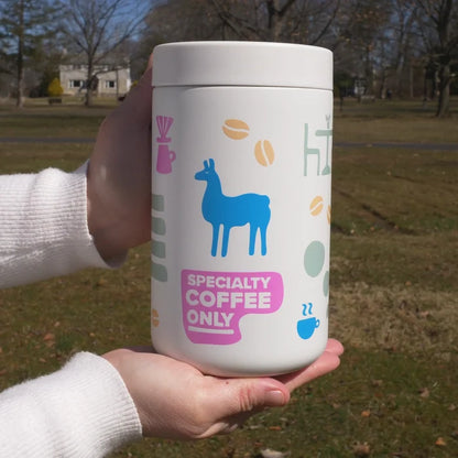 Person holding a white coffee canister with colorful designs and text in an outdoor setting