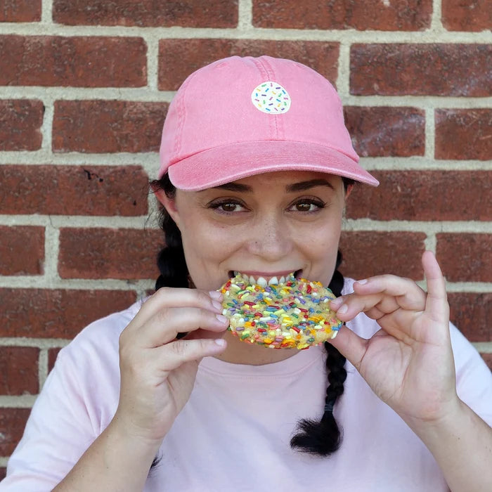 Image of woman in coral dad hat with embroidered cookie hat biting a sprinkle sugar cookie