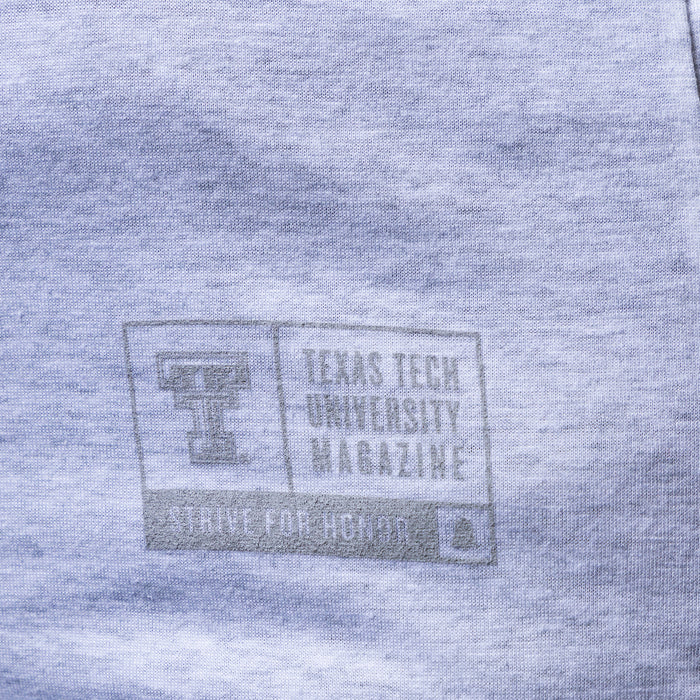 Close-up of a heather gray fabric surface with a faded gray printed Texas Tech University Magazine logo and text.