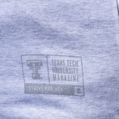 Close-up of a heather gray fabric surface with a faded gray printed Texas Tech University Magazine logo and text.