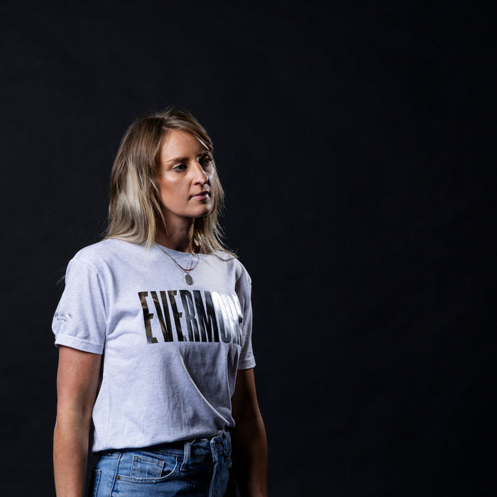 A woman with blonde hair wearing a light gray graphic t-shirt with the word 'EVERMIORE' printed in black across the chest, paired with blue jeans, set against a solid black background.