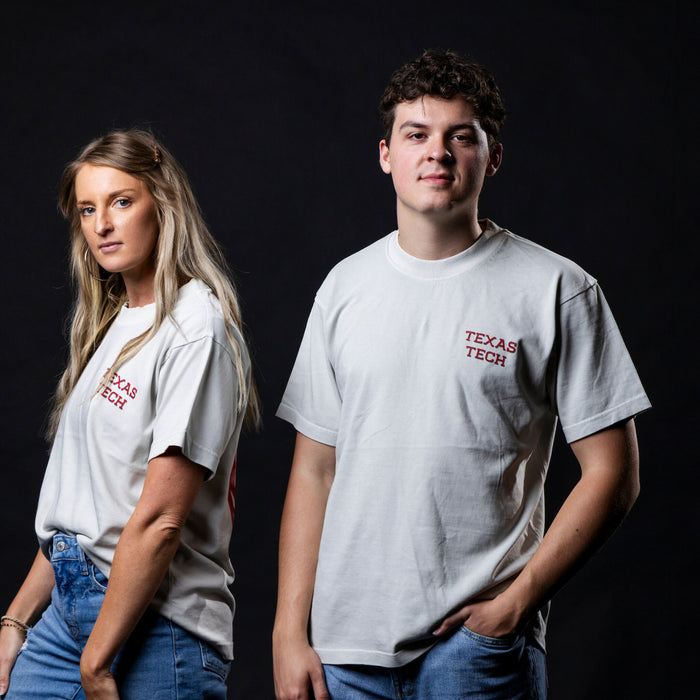 A man and a woman wearing matching white Texas Tech t-shirts with red text on the left chest, posed against a dark studio background.